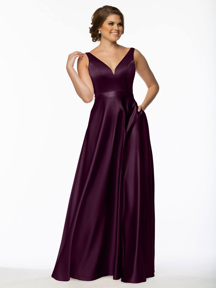 A woman smiles in the Avery Austin 80118, a sleeveless, floor-length satin bridesmaid dress with pockets and a V-neckline, posing with one hand on her hip and the other touching her hair against a plain white background.