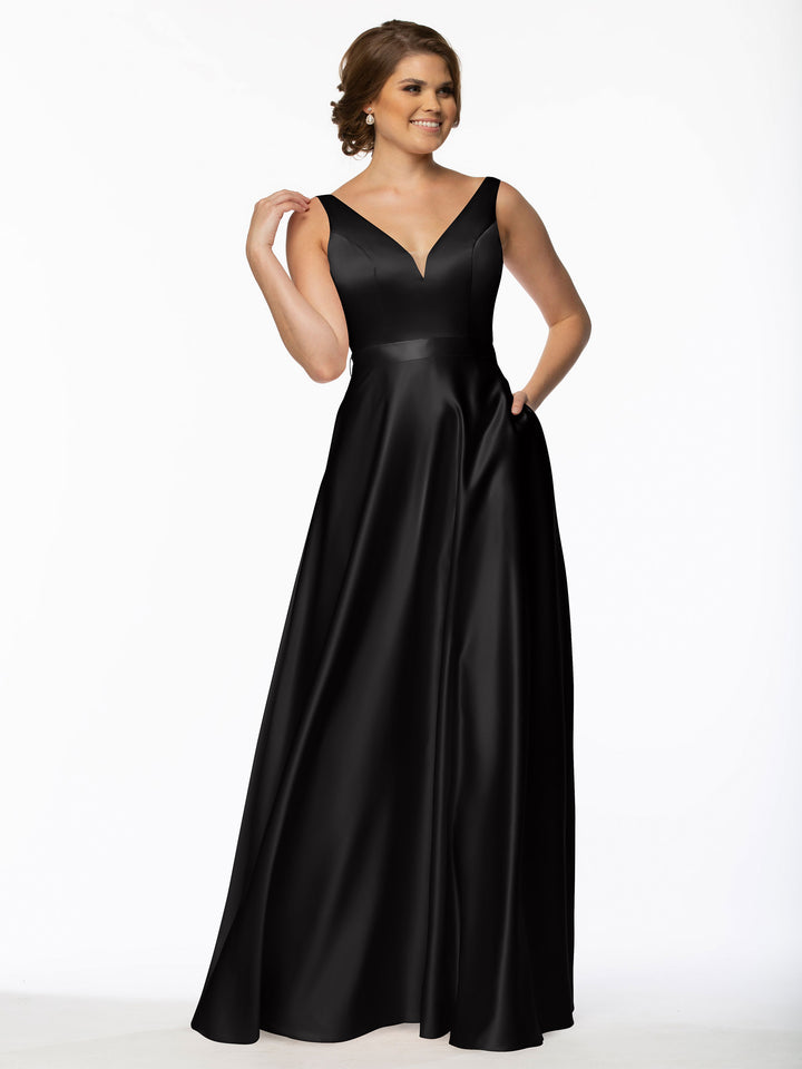A woman in a black Avery Austin long satin bridesmaid dress (style 80118) with a V-neckline stands against a white background, smiling and touching her hair with one hand as the other rests on her hip.