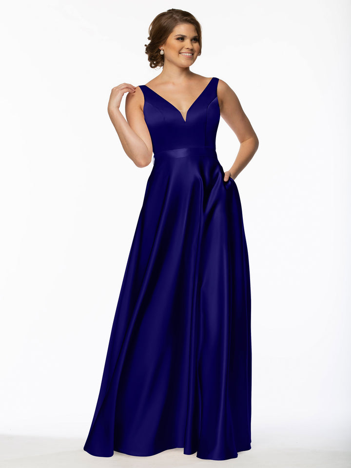 A woman stands smiling against a plain white background, wearing Avery Austin’s 80118: a sleeveless, floor-length royal blue satin bridesmaid dress with a V-neckline.