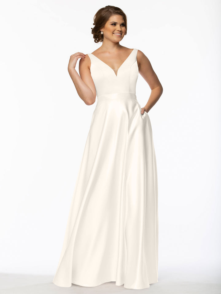 A woman stands smiling against a plain white background, wearing Avery Austin’s 80118—a sleeveless, floor-length bridesmaid dress with a deep V-neckline and fitted bodice. She raises one hand near her hair as the skirt flows elegantly.