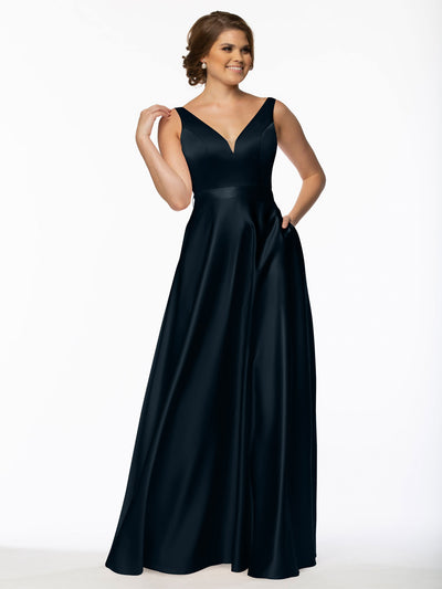A woman smiles against a white background, wearing Avery Austin’s 80118: a sleeveless, floor-length black satin bridesmaid dress with pockets and a V-neckline. She touches her hair with one hand and rests the other in her pocket.