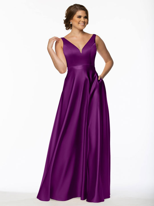 A woman stands smiling, one hand touching her hair, wearing the Avery Austin 80118 purplish satin bridesmaid dress with a floor-length, sleeveless design and V-neckline against a plain white background.