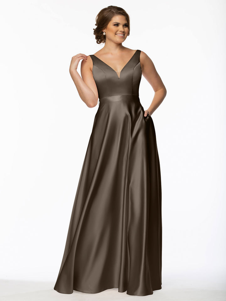 A woman stands against a plain white background, smiling and touching her hair while wearing Avery Austin’s 80118: a floor-length satin bridesmaid dress with sleeveless V-neckline, fitted bodice, and full skirt.
