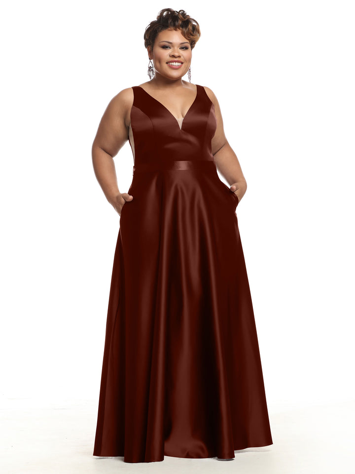 A smiling woman wears Avery Austin’s 80118 burgundy satin bridesmaid dress with a deep V-neckline, pockets, and sleeveless floor-length design, standing against a plain white background.