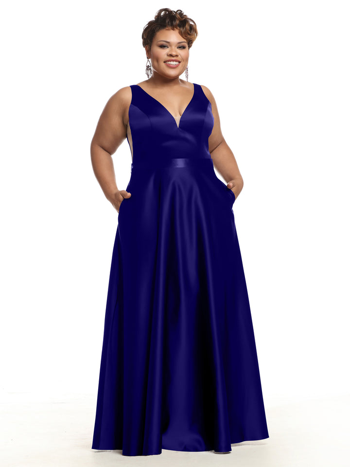 A woman stands and smiles against a plain white background, wearing Avery Austin’s 80118 sleeveless deep blue satin bridesmaid dress with pockets and a V-neckline.