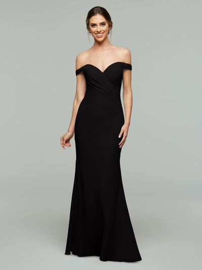 A woman wearing Avery Austin’s 80122 off-the-shoulder, floor-length black bridesmaid dress stands smiling against a plain gray background.