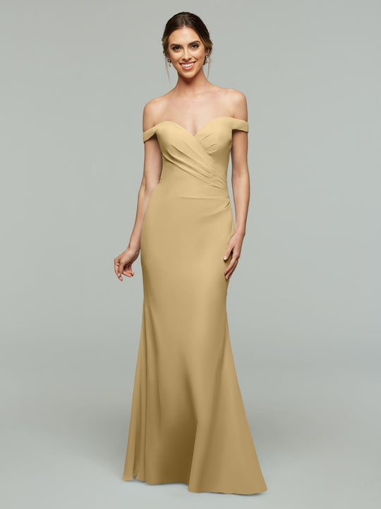 A woman wearing Avery Austin's 80122 floor-length bridesmaid dress in champagne stands against a plain grey background, smiling with her hair in an updo. The off-the-shoulder gown blends elegance and modern style.
