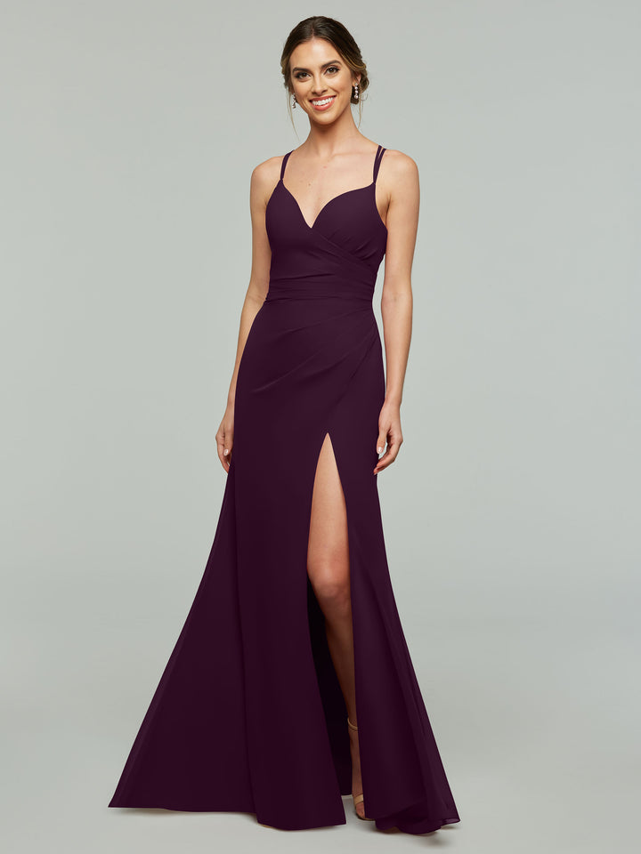 A woman wearing the Avery Austin 80130 dress in Aubergine, a sleeveless v-neck chiffon bridesmaid gown with spaghetti straps and a high slit, stands against a plain light gray background, smiling at the camera.
