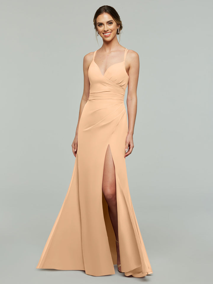 A woman smiles against a gray background in Avery Austin’s blush sleeveless sheath bridesmaid dress (style 80130) featuring spaghetti straps, a V-neckline, and a high slit.