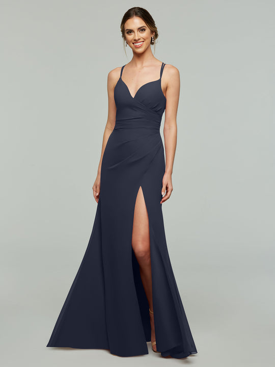 A woman stands against a light gray background wearing Avery Austin style 80130, a floor-length navy blue sheath bridesmaid dress with spaghetti straps, sweetheart neckline, and a high side slit.