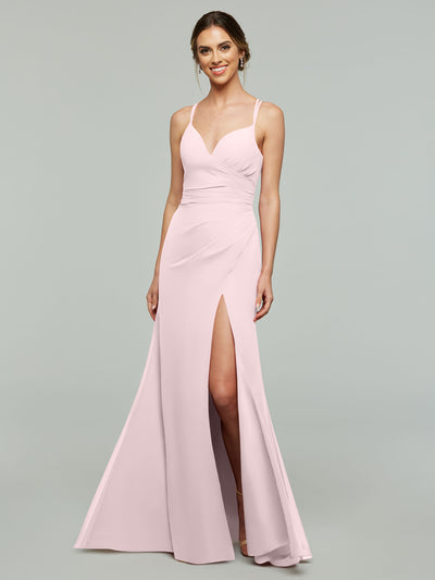 A woman stands smiling against a plain gray background wearing Avery Austin style 80130, a light pink, floor-length chiffon bridesmaid dress with thin straps, a v-neckline, fitted bodice, and thigh-high slit.