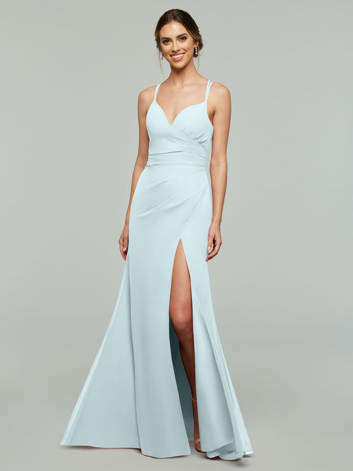 A woman smiles against a gray background, wearing Avery Austin style 80130—a light blue v-neck chiffon bridesmaid dress with a fitted bodice, side ruching, and high slit.