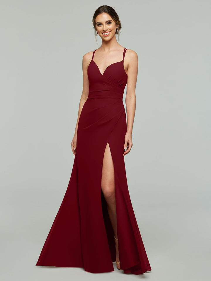 A woman wears the Avery Austin TH-80130 bridesmaid dress in wine, featuring a v-neck, thin straps, fitted bodice, and thigh-high slit. She smiles while standing against a plain light gray background.