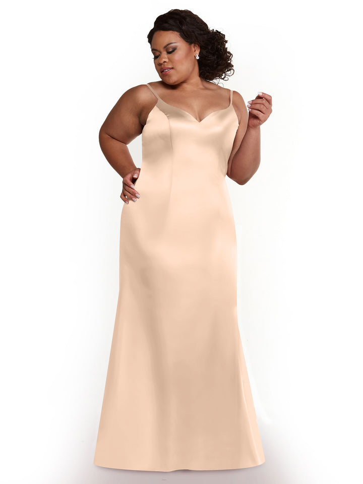 A woman models Avery Austin style 80132, a long champagne fit-and-flare dress with thin straps and sleeveless design. She poses against a white background, dark curly hair pulled back, wearing earrings and striking an elegant pose.