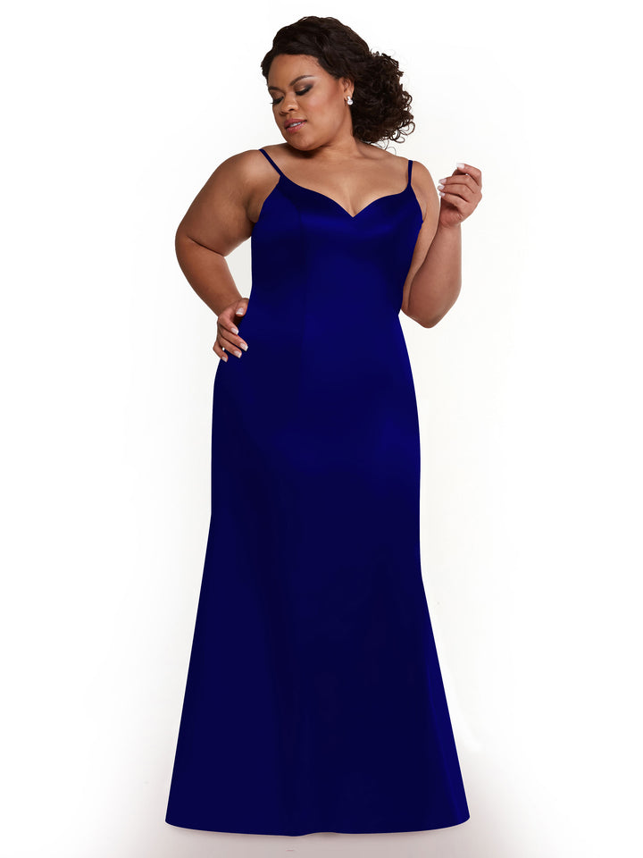A woman wears the Avery Austin 80132: a floor-length, sleeveless, deep blue fit-and-flare dress with thin straps. She stands against a white background, looking down and smiling slightly, one hand on her hip, the other raised.