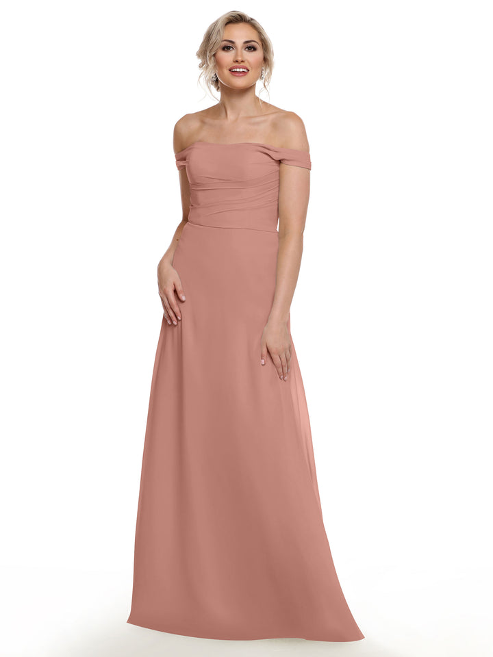A woman in the Avery Austin 80136 blush pink, off-the-shoulder chiffon dress stands against a white background, smiling with one hand by her side and the other on her waist.
