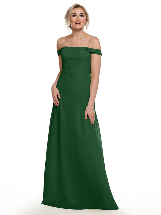 A woman models Avery Austin style 80136: a dark green, floor-length off-the-shoulder chiffon dress, standing against a white background and smiling with one hand at her side and the other resting on her hip.