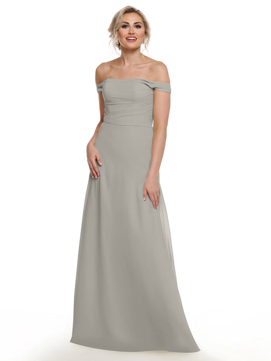A woman models Avery Austin’s 80136, a light gray off-the-shoulder chiffon dress, standing and smiling in front of a white background with her hand by her side.