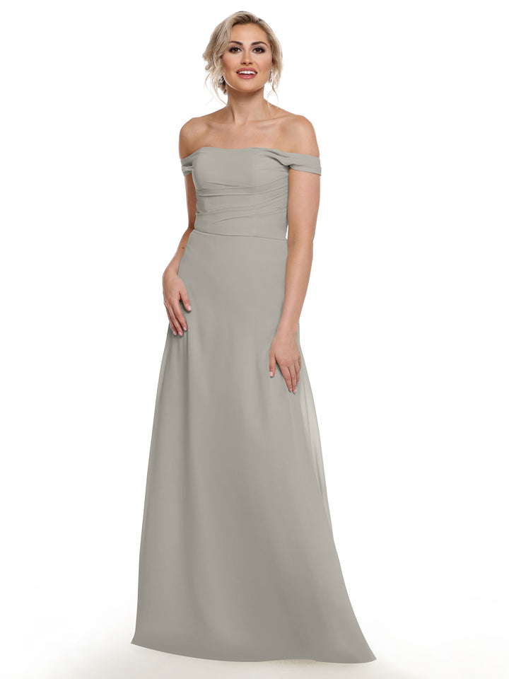 A woman models Avery Austin’s 80136, a light gray off-the-shoulder chiffon dress, standing and smiling in front of a white background with her hand by her side.
