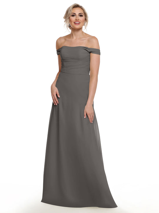 A woman in Avery Austin’s 80136 off-the-shoulder, floor-length chiffon dress stands smiling against a plain white background, her hair up, one hand by her side and the other gently touching her waist.