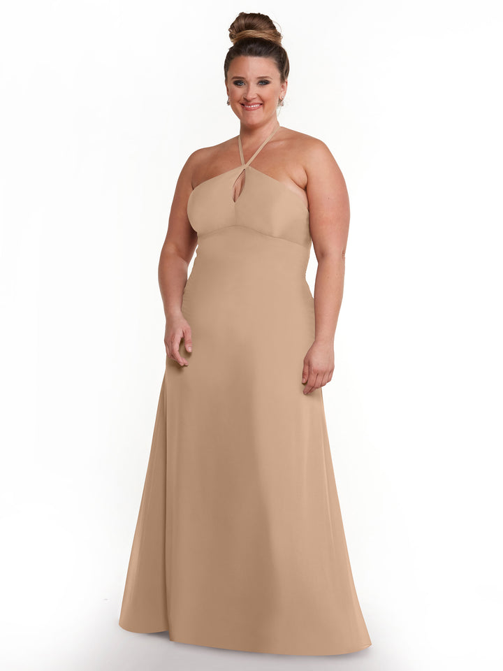 A woman with light skin and brown hair in a high bun smiles while modeling Avery Austin’s 80137 sleeveless floor-length beige halter dress with a keyhole back, standing against a plain white background.