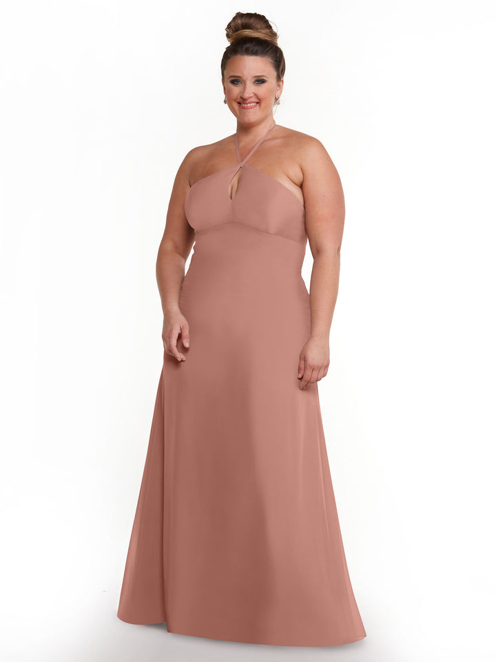 A woman with brown hair in a bun models Avery Austin’s minimalistic blush pink floor-length gown, style 80137, featuring a keyhole back, as she smiles against a plain white background.