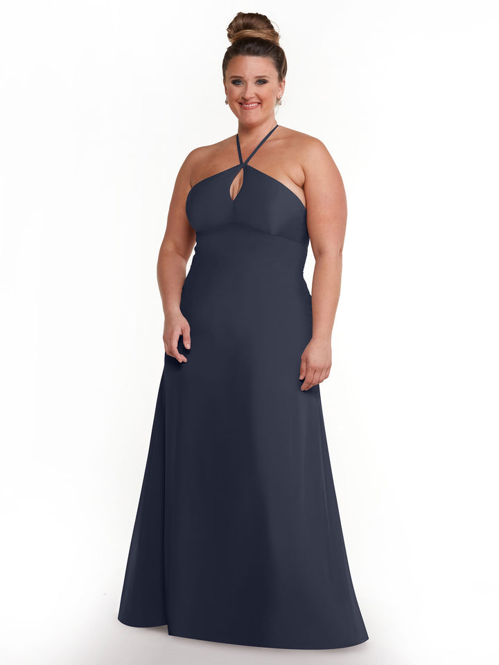 A woman with light skin and brown hair in a bun models the Avery Austin 80137, a dark navy, floor-length halter dress with a keyhole neckline and minimalistic design, standing and smiling against a plain white background.