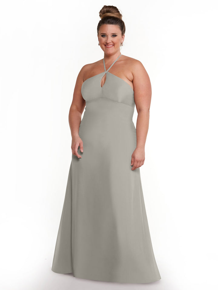 A woman with her hair in a bun is wearing Avery Austin’s 80137, a long light gray halter dress with a keyhole cutout, while standing and smiling against a plain white background.