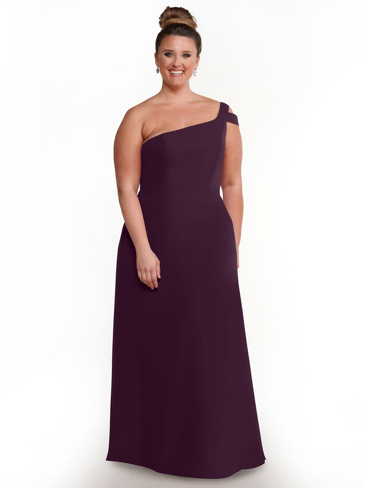 A woman stands smiling against a white background in Avery Austin’s aubergine one-shoulder floor-length dress (style 80139), her hair in a high bun and drop earrings completing the elegant look.