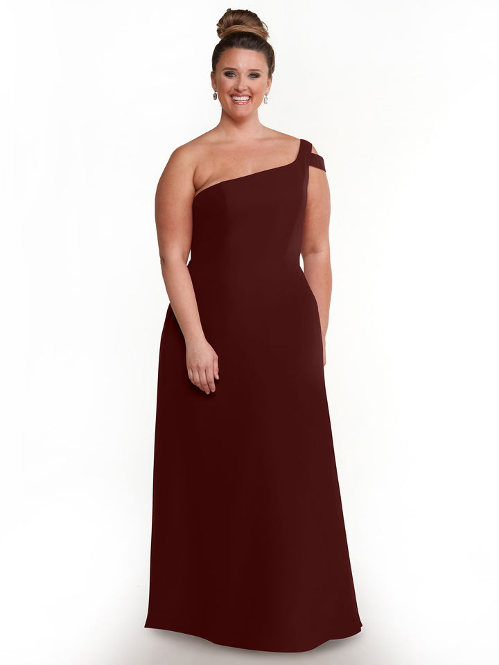 A woman smiles in a floor-length, merlot one-shoulder Avery Austin 80139 dress with a chic side slit. Her hair is in a bun, and she wears drop earrings against a plain white background.