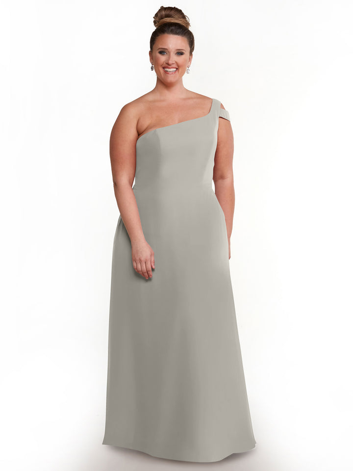 A woman smiles in Avery Austin style 80139, a chic light gray one-shoulder dress featuring a shoulder cutout. She wears her hair in a neat updo with earrings, posing against a plain white background.