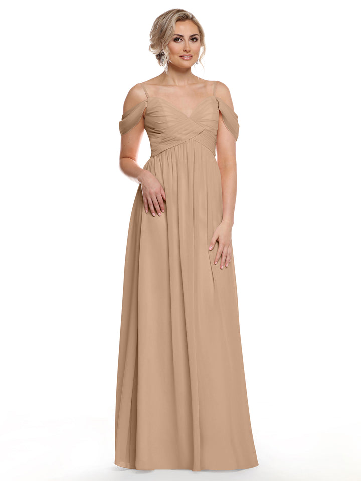 A woman models Avery Austin style 80142, a long buff chiffon cold shoulder dress with a pleated bodice and sheer straps. Her blonde hair is styled up and she pairs the look with statement earrings against a white backdrop.