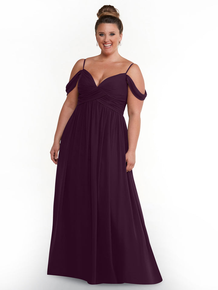 A woman stands smiling in Avery Austin’s 80142 floor-length aubergine chiffon dress, featuring off-the-shoulder draped sleeves, a pleated bodice, and spaghetti straps, against a plain white background.