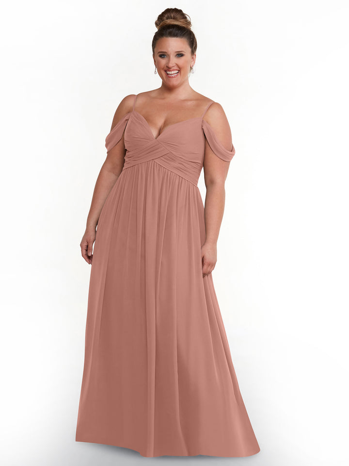 A woman wears the Avery Austin 80142, a blush pink off-the-shoulder chiffon dress. She stands smiling against a white background with her hair in a high bun and drop earrings.