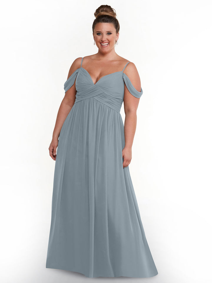 A smiling woman with her hair in a bun wears Avery Austin style 80142, a long light blue chiffon cold shoulder dress with a gathered bodice, standing against a plain white background.