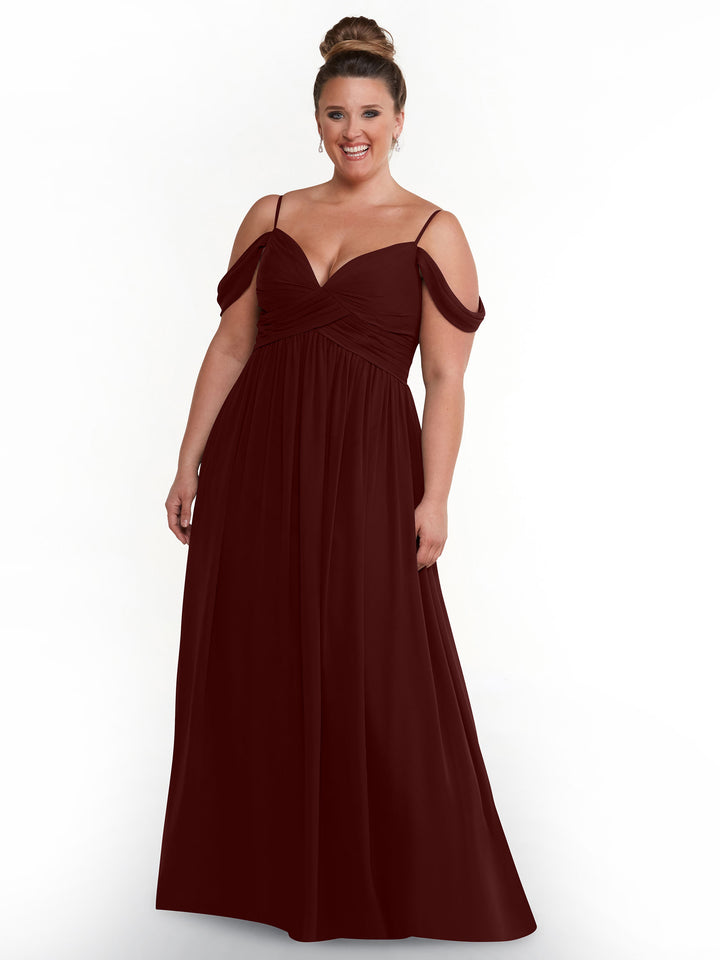 A smiling woman with her hair in a bun wears the Avery Austin 80142 floor-length dress in Merlot, featuring a boho chic style with an off-the-shoulder sweetheart neckline and pleated bodice, standing against a plain white background.