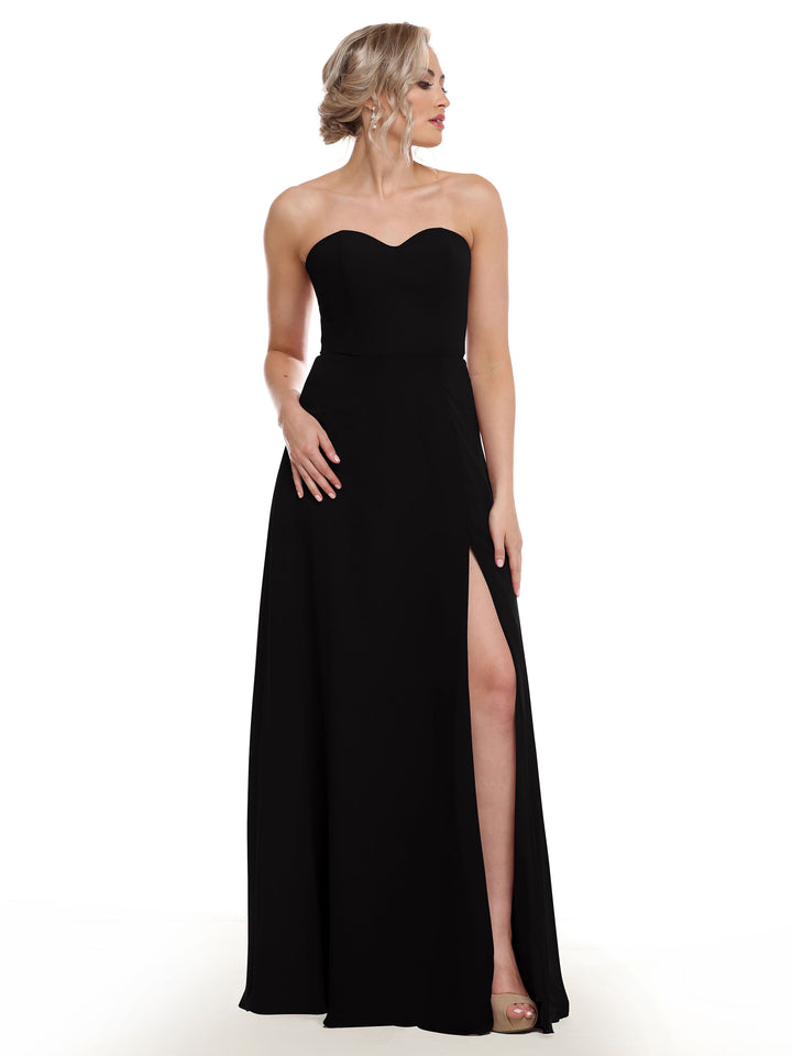 A woman stands against a plain white background wearing Avery Austin’s 80143: a black, floor-length strapless sweetheart gown with a sash back and side slit.