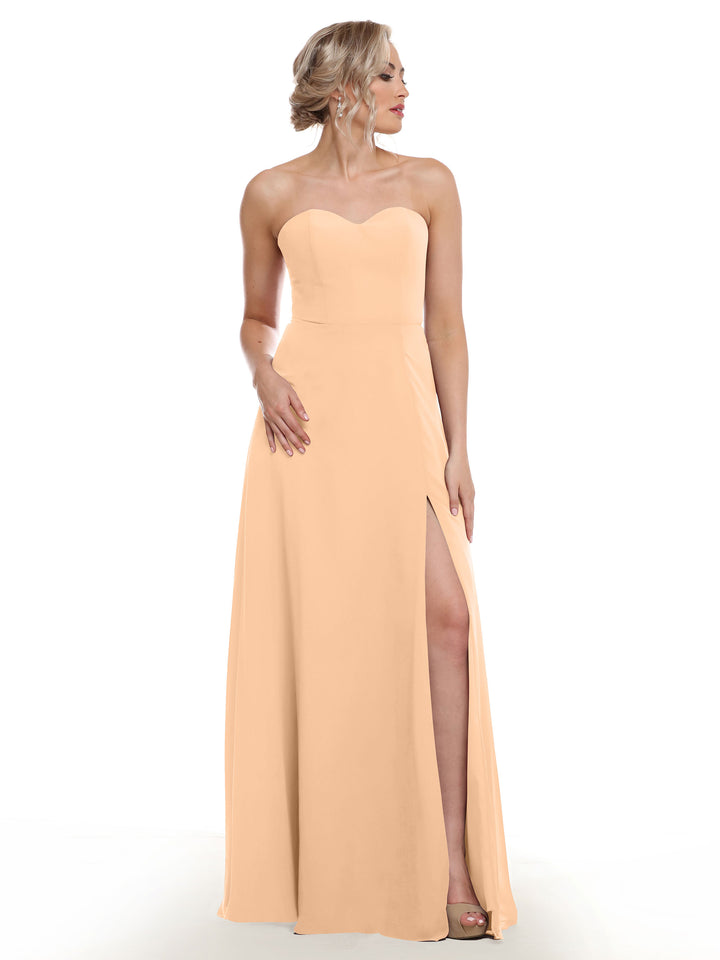 A woman models the Avery Austin 80143, a strapless sweetheart dress in peach with sash back and side slit, standing and looking left against a white background.