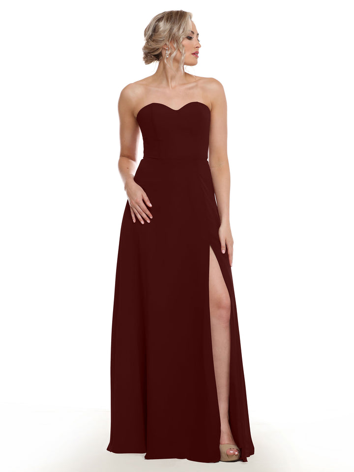 A woman models Avery Austin’s 80143: a dark red, strapless sweetheart gown with a floor-length skirt, sash back, and high side slit, standing against a white background and gazing to her left.