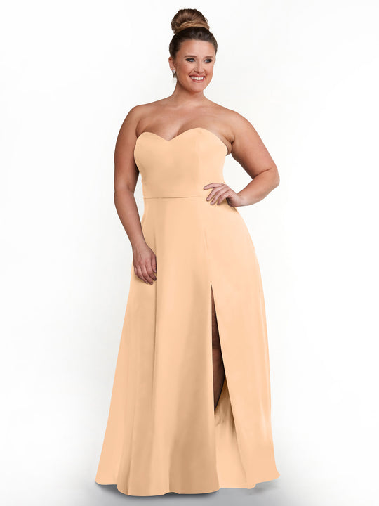 A woman with brown hair in a bun wears Avery Austin’s 80143 strapless sweetheart light peach dress with a side slit and thigh-high cut, standing and smiling against a plain white background.