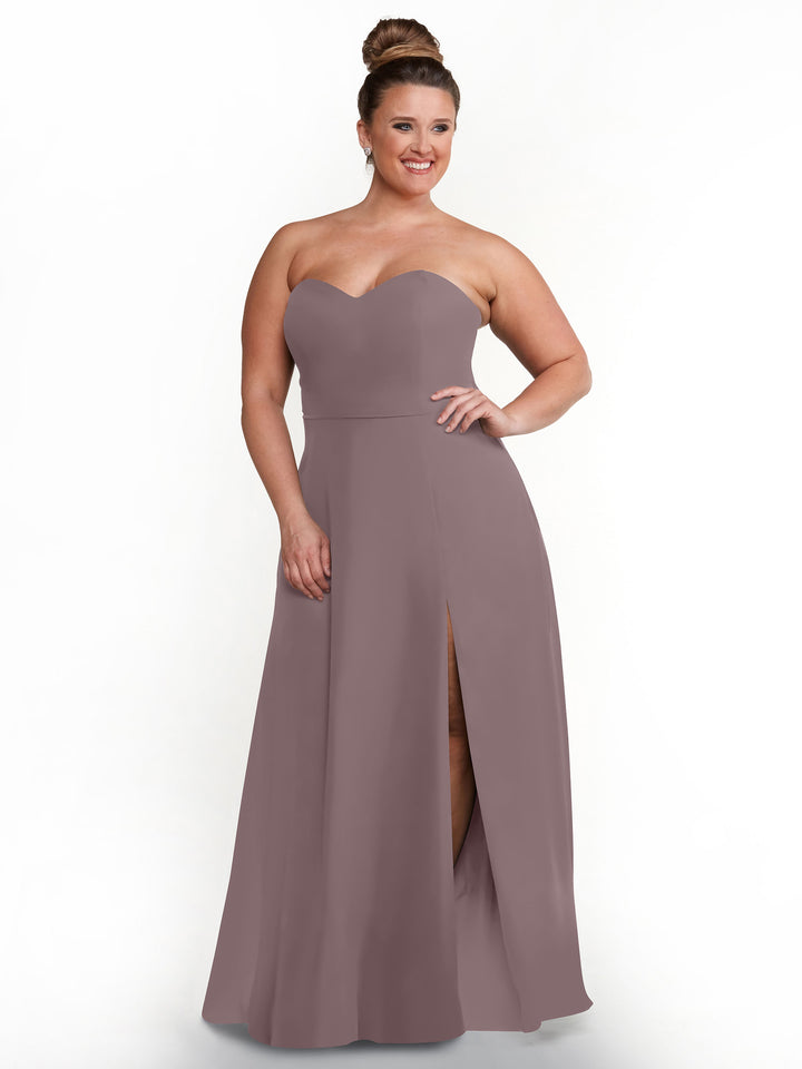 A woman smiles with one hand on her hip, modeling Avery Austin style 80143—a mauve strapless sweetheart dress with a floor-length skirt, side slit, and elegant sash back—against a white background.