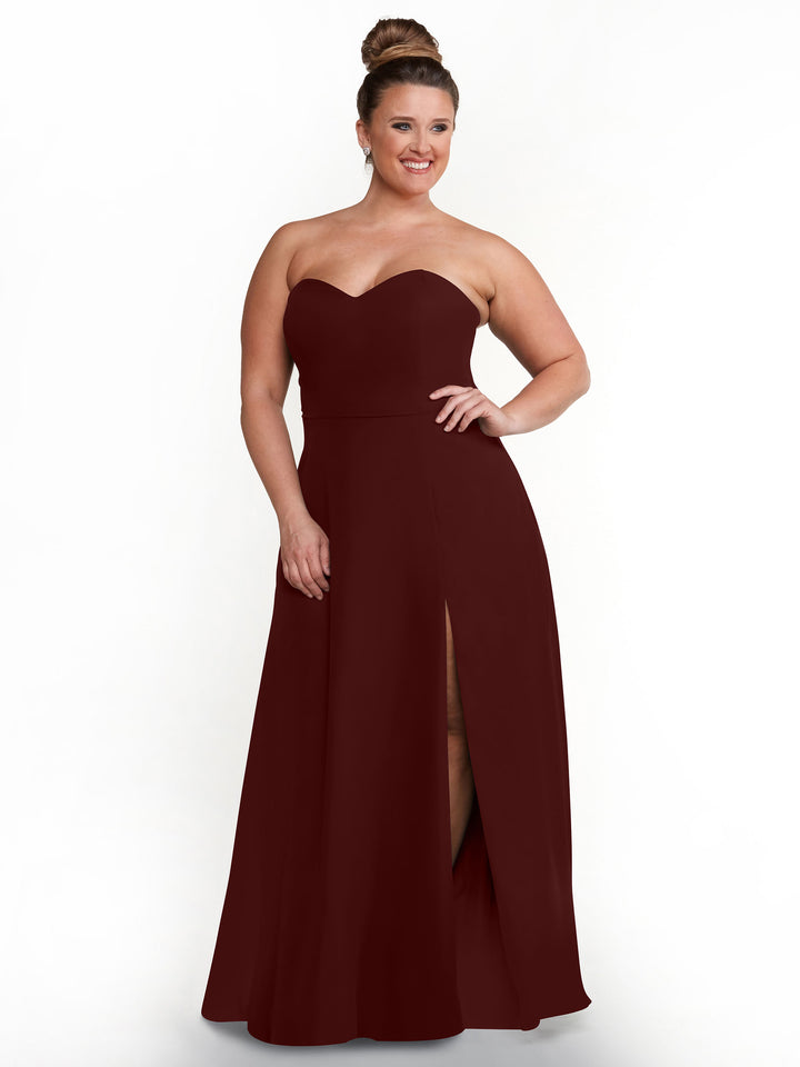 A woman with her hair in a bun smiles as she models the Avery Austin 80143 dress—a burgundy, strapless sweetheart gown with a sash back and floor-length skirt featuring a high side slit—against a plain white background.