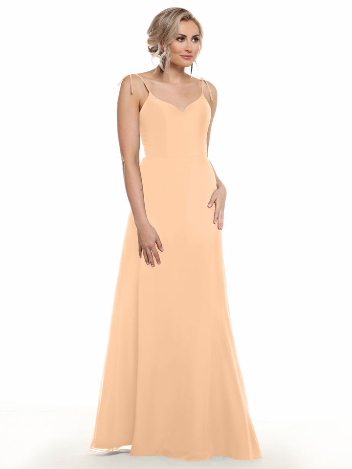 A woman with short blonde hair and a neutral expression models Avery Austin’s 80144: a long, peach flowy dress with thin straps and a V-neck, standing against a white background with one hand on her hip.