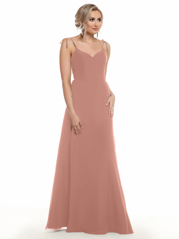 A woman with short, wavy blonde hair and a neutral expression models the Avery Austin 80144: a sleeveless, floor-length blush pink flowy dress featuring thin shoulder straps and a subtle V-neck, set against a white background.
