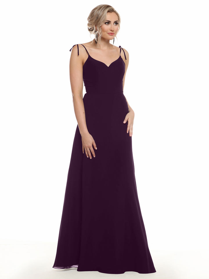 A woman with blonde, wavy hair styled up poses with one hand on her hip, wearing Avery Austin’s floor-length chiffon dress (80144) in deep purple, featuring a V-neck and thin shoulder straps against a white background.