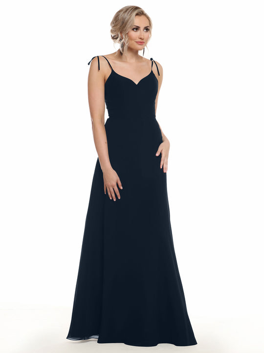 A woman with blonde hair in an updo wears Avery Austin’s 80144, a long black chiffon dress with thin straps and a fitted bodice, standing against a white background.