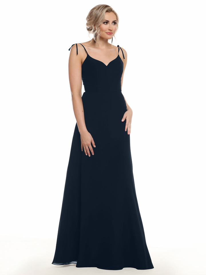 A woman with blonde hair in an updo wears Avery Austin’s 80144, a long black chiffon dress with thin straps and a fitted bodice, standing against a white background.