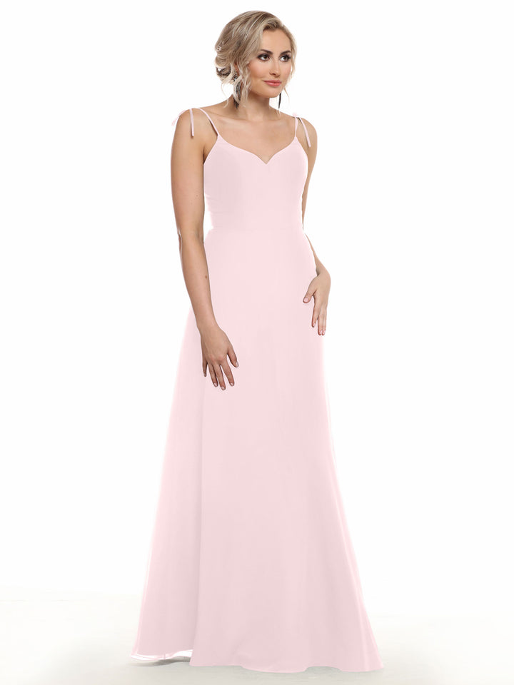 A woman with blonde, wavy hair styled up wears Avery Austin 80144—a long, light pink chiffon dress with thin straps and a V-neck—as she stands against a plain white background, gazing slightly to the side.