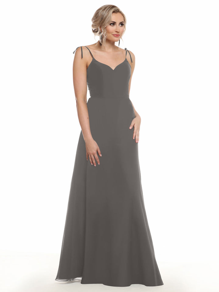 A woman with blonde hair in a loose updo wears Avery Austin’s 80144, a long, sleeveless dark gray chiffon dress with thin shoulder ties, posed against a plain white background.