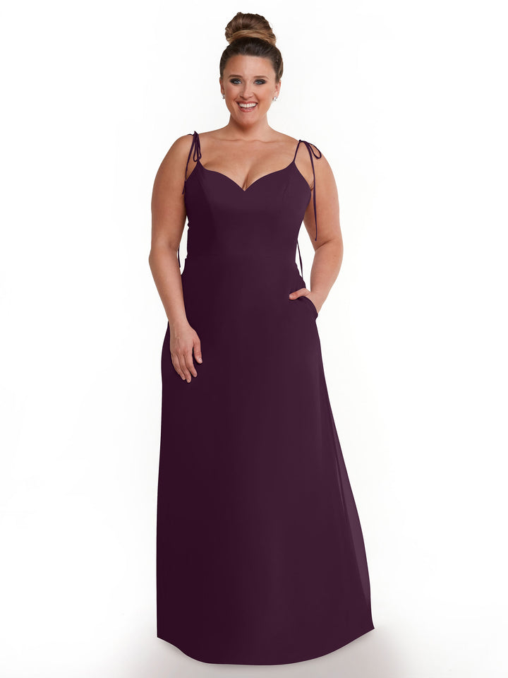 A woman with her hair in a bun models Avery Austin’s 80144: a sleeveless, floor-length deep purple chiffon dress with thin straps, V-neck, and pockets, posing against a plain white background.